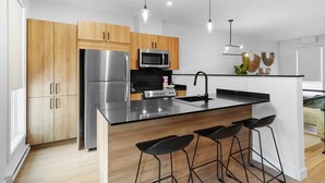 Una cocina moderna con electrodomésticos de acero inoxidable, armarios de madera, taburetes de bar negros e iluminación colgante.
