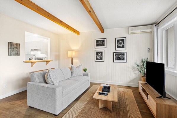 Un salon avec un canapé gris, une table basse en bois, un téléviseur à écran plat et des œuvres d’art florales encadrées accrochées au mur.