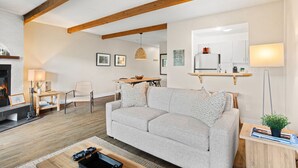 A modern living room with a grey sofa, wooden beams, a fireplace, a dining area, and a kitchen with a mirror and shelf. - Stunning One-Bedroom Condo With Lake View (Mont-Tremblant)