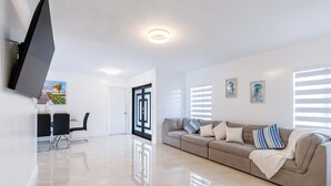 A modern living room with a sofa, a flat-screen TV mounted on the wall, and a dining area with chairs and a table. - Escape to Miami/Heather+Pool/9pl/BBQ/pool table. (Miami)