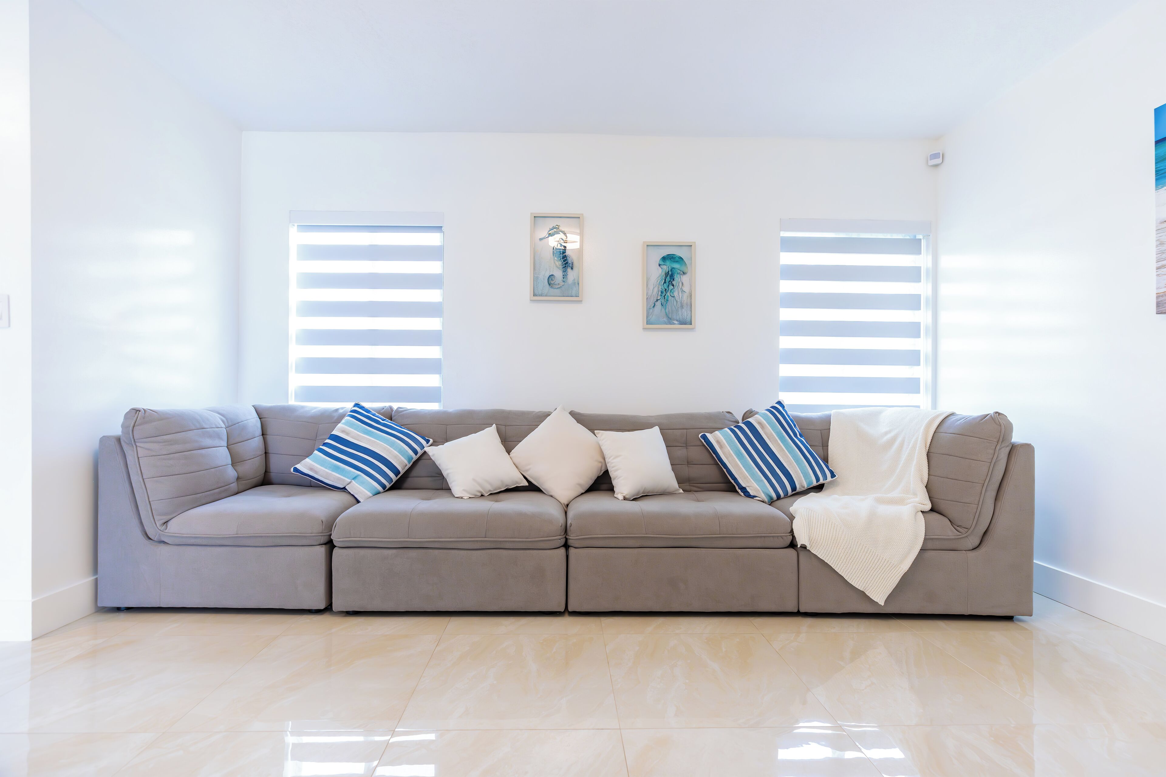 A modern living room with a beige sectional sofa, two wall-mounted pictures, and a large window with blinds.