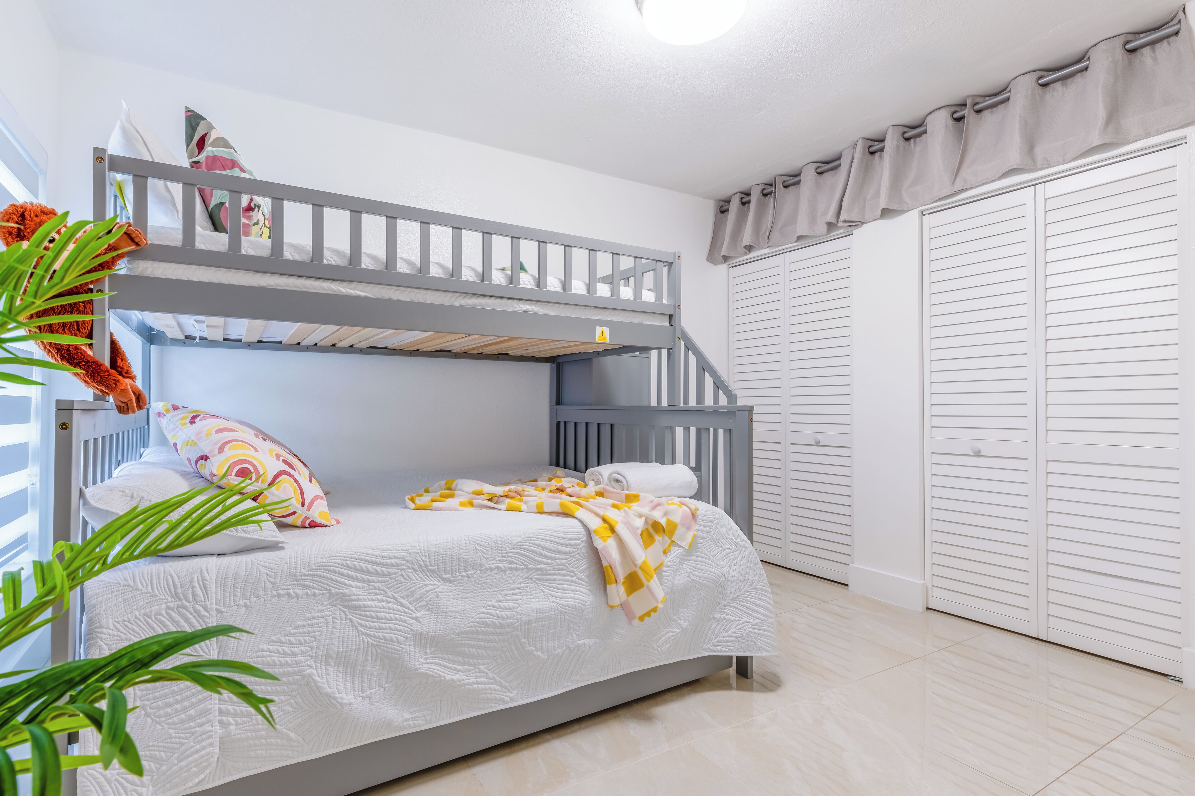 A bunk bed with a white mattress and a checkered blanket, a small bedside table with a lamp, and a window with curtains.