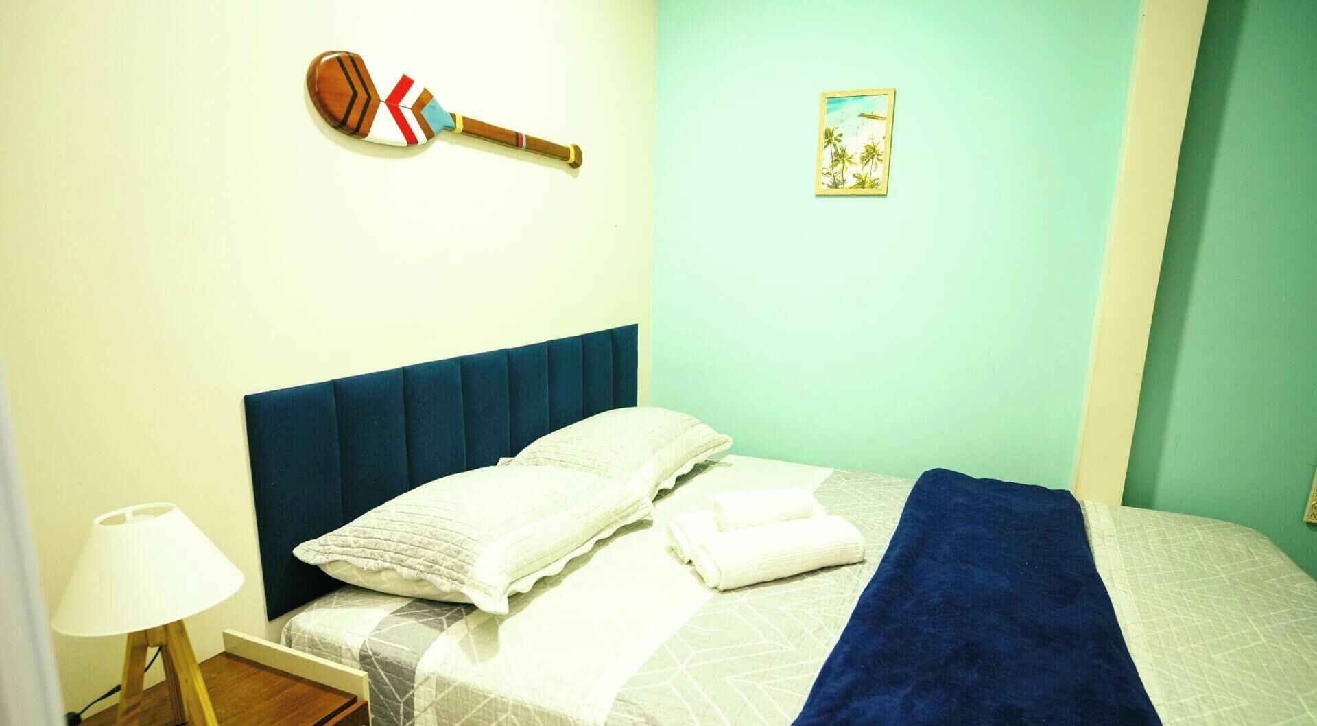 A bedroom with a bed, pillows, a blue headboard, a lamp, a picture frame, and a wall-mounted object.
