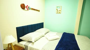 A bedroom with a bed, pillows, a blue headboard, a lamp, a picture frame, and a wall-mounted object.