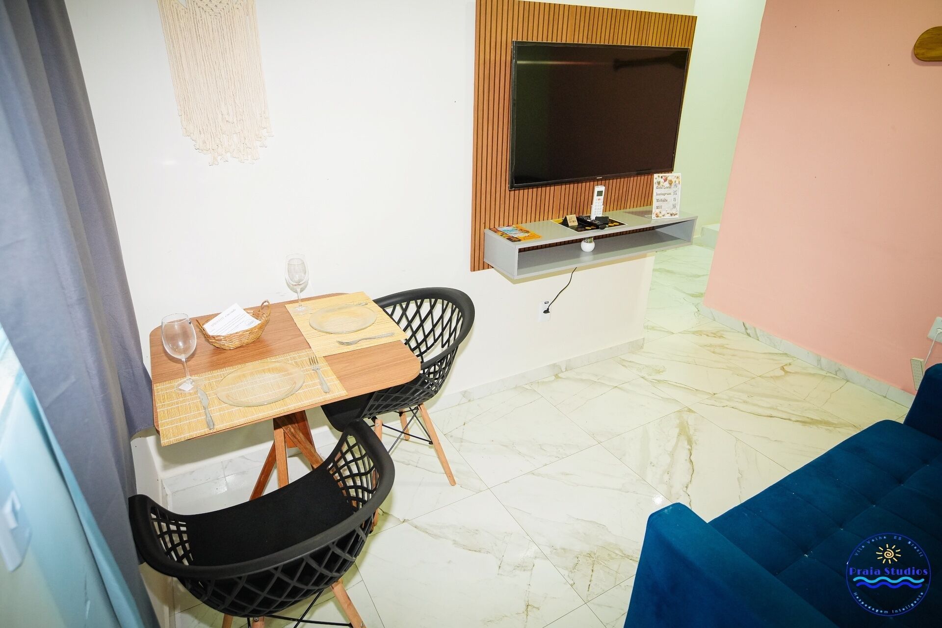 A dining area with a table set for two, a TV mounted on the wall, and a blue sofa in the background.
