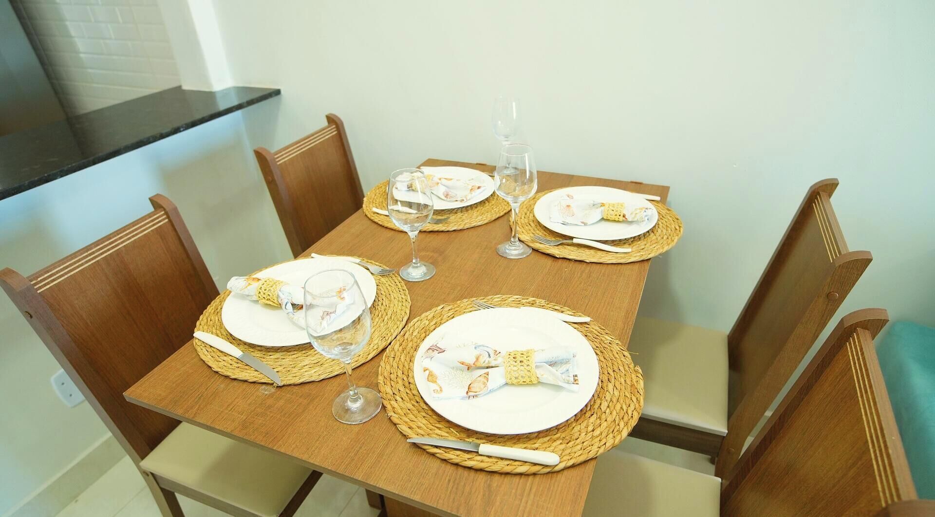 Una mesa de comedor preparada para cuatro personas, con individuales, platos, cubiertos y copas de vino.