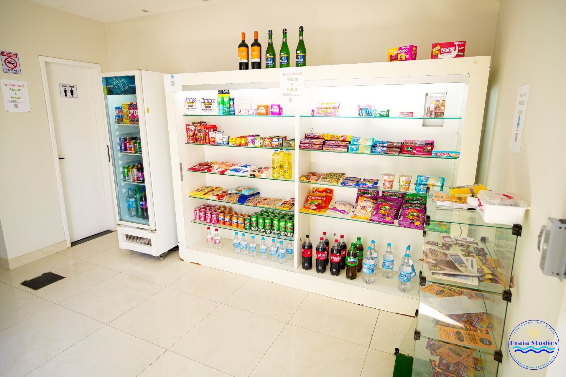 Una habitación con un refrigerador lleno de bebidas y aperitivos, una puerta con un letrero de 'no fumar' y una vitrina de vidrio con periódicos.