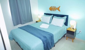 A neatly made bed with a blue bedspread and pillows, a wooden fish wall decoration, and bedside lamps on a nightstand. - Praia Studios (Vila Velha)