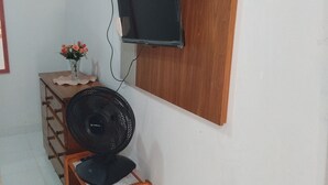 Una habitación con una televisión montada en un panel de madera, un ventilador de pie, un mueble de madera con cajones y un florero con flores sobre una mesita.
