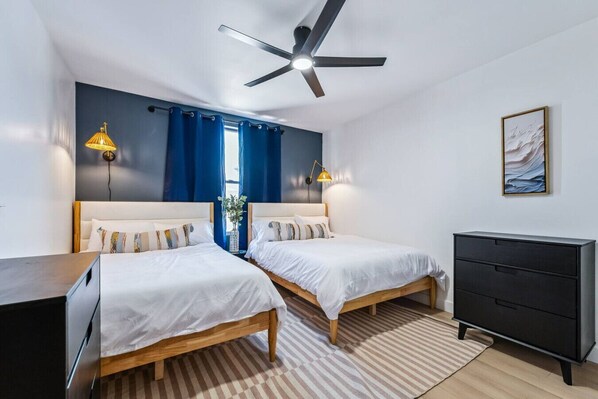 A double room with two beds, a ceiling fan, a dresser, and a painting on the wall.
