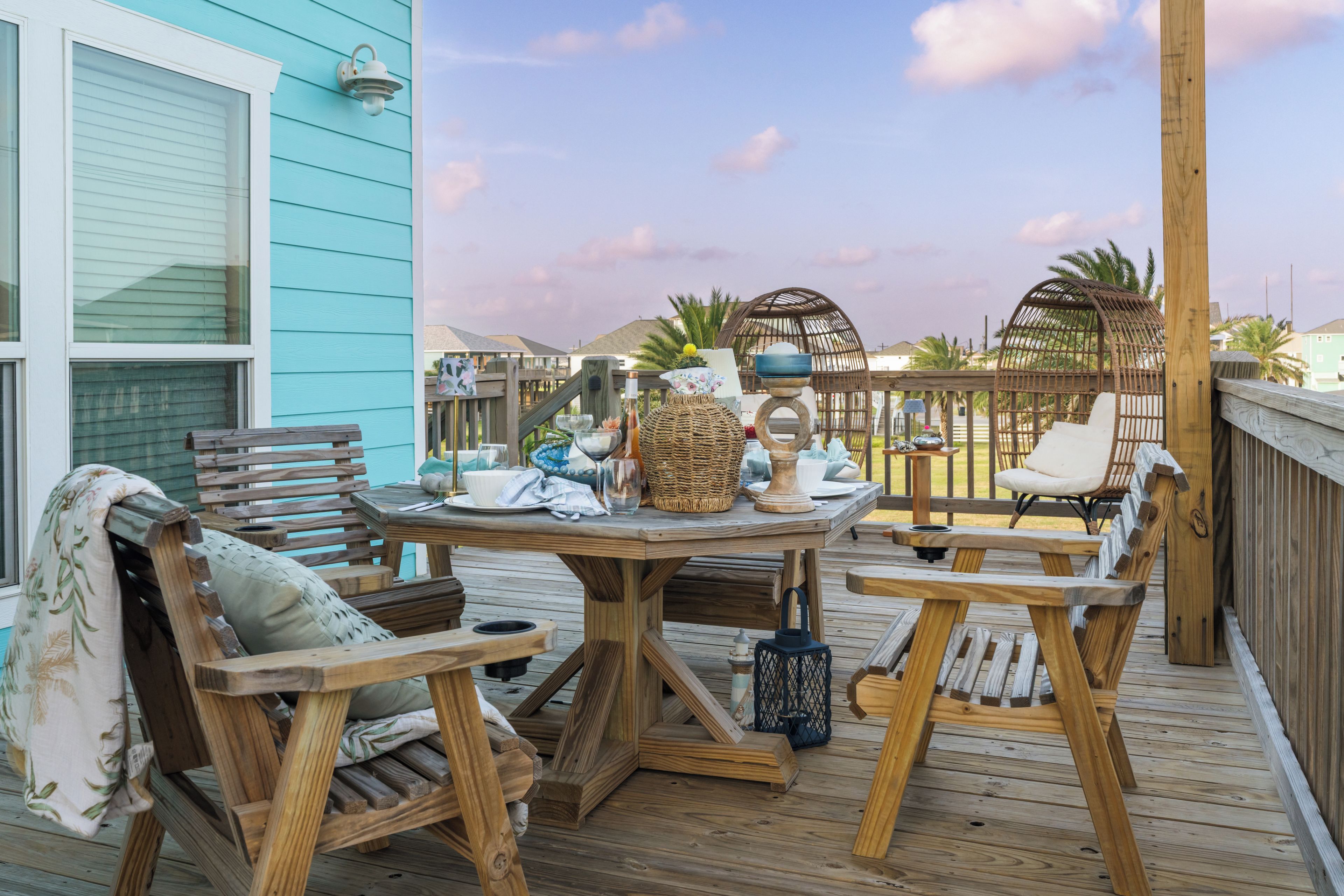 An outdoor wooden deck with a table set for dining, surrounded by wicker chairs and a wooden bench.