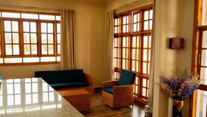 A room with a wooden floor, a green sofa, wicker chairs, and large windows with curtains. - INOGA LODGE KARATU (Karatu)