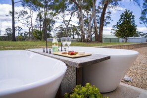 Ein Wellnessbereich mit zwei großen weißen Badewannen, einem Holztisch mit einer Flasche, Gläsern und Snacks, umgeben von Bäumen und blauem Himmel.