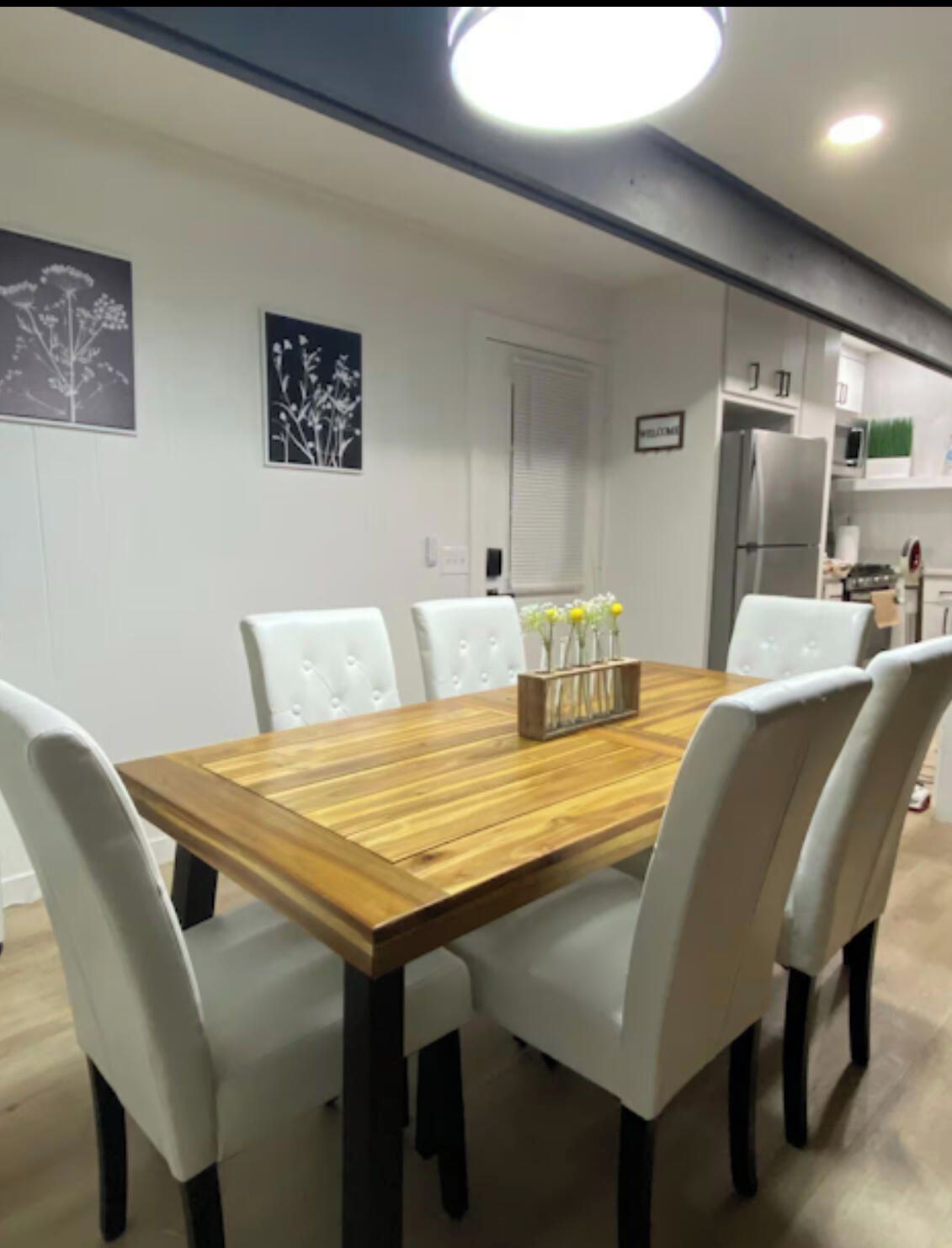 A dining area with a wooden table, white upholstered chairs, and a vase with yellow flowers.