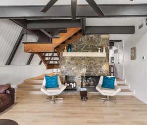 A modern living room with a stone fireplace, two white chairs with blue cushions, a wooden staircase, and a kitchen in the background. - Pet-Friendly Lakehouse w/ Gameroom & Kayaks (Granbury)