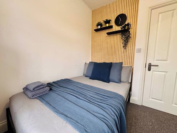 Een net opgemaakt bed met een blauwe deken en kussens, een wandklok en een deur aan de rechterkant.