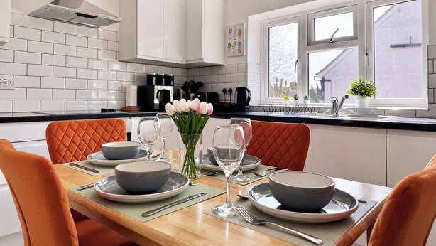 Una cocina moderna con armarios blancos, encimera negra y zona de comedor con sillas anaranjadas y una mesa de madera para cuatro personas.