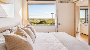 Une chambre avec une grande fenêtre, un lit avec une literie de couleur beige et une vue sur la verdure.