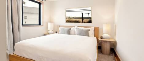 Un dormitorio ordenado con una cama grande, dos mesitas de noche con lámparas, una ventana con cortinas y un cuadro enmarcado en la pared.