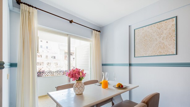 Un coin repas comprenant une table, des chaises, un vase avec des fleurs, un pichet en verre et une assiette de fruits.