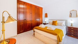 Une chambre avec une grande armoire en bois, un lit recouvert d’une courtepointe blanche et jaune moutarde, une table de chevet avec une lampe et un petit miroir accroché au mur.