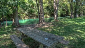 A picnic table in a wooded area with a river in the background. - 4-bedroom house in Utopia with 450’ of private crystal clear Sabinal river front (Utopia)