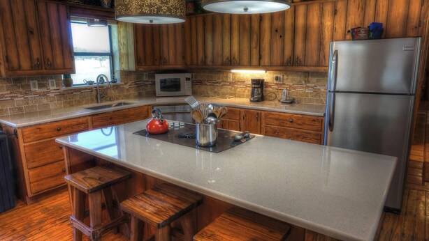 A kitchen with wooden cabinets, a stainless steel refrigerator, a microwave, and a coffee maker. There are wooden stools and a red kettle on the counter. - 4-bedroom house in Utopia with 450’ of private crystal clear Sabinal river front (Utopia)