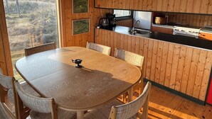 Un intérieur en bois comprenant une table à manger, des chaises, un luminaire suspendu, un coin cuisine avec une cuisinière et une fenêtre donnant sur l’extérieur.