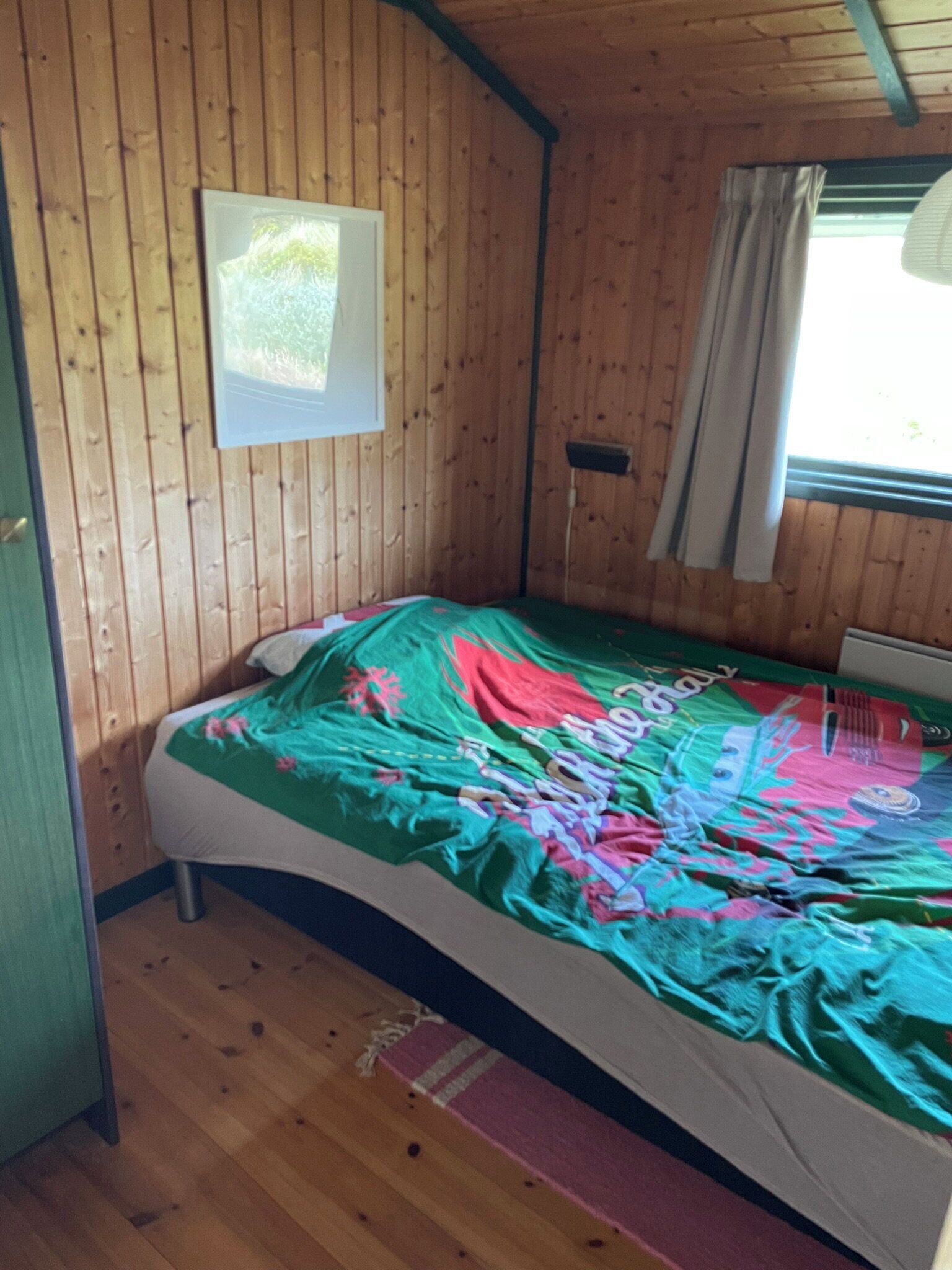 Een houten kamer met een bed, een raam met gordijnen en een aan de muur hangend schilderij.