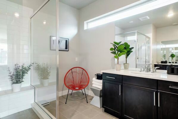 A modern bathroom with a glass shower enclosure, a red chair, a large mirror, and dark cabinets.