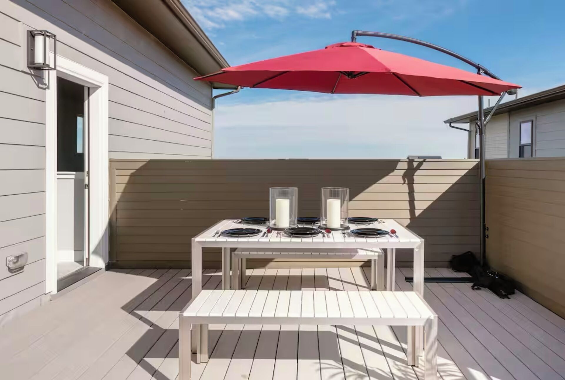 Een terras buiten met een witte tafel en bank, een rode parasol en uitzicht op de lucht.