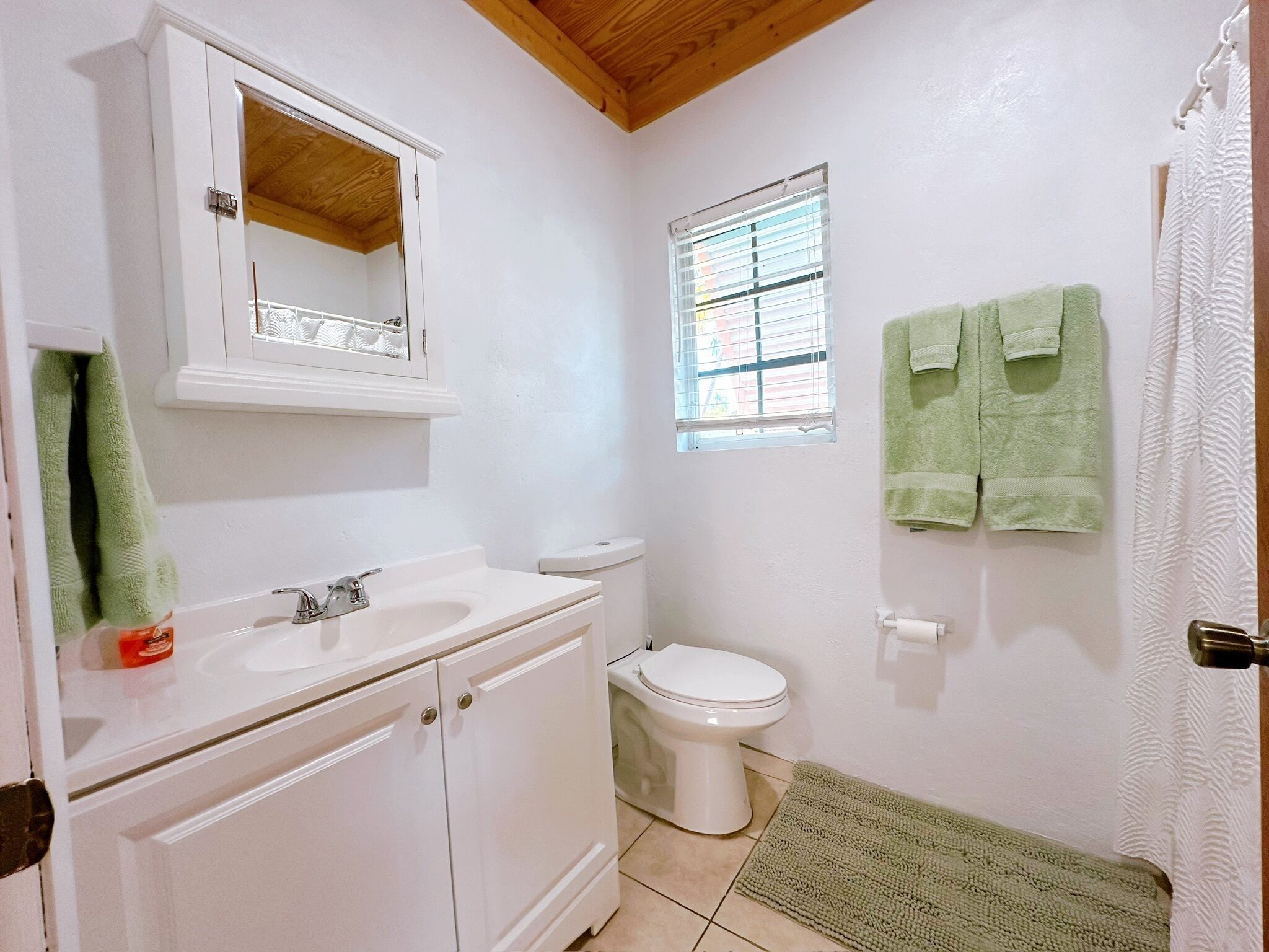 Une salle de bain avec un lavabo blanc, un miroir et des toilettes. Il y a une fenêtre avec des stores, une serviette verte et un rideau de douche.