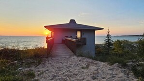 Ein Strandhaus mit einer Holzterrasse, die zu einer überdachten Veranda führt und auf den Ozean im Abendrot blickt.