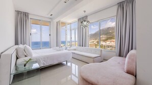 Ein modernes Schlafzimmer mit Bett, Sofa, Sessel und großen Fenstern mit Blick auf das Meer und die Berge.