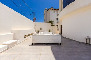 A rooftop terrace with a white sofa and a small table, surrounded by white walls and a clear blue sky.