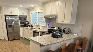 A modern kitchen with white cabinets, a stainless steel refrigerator, a built-in oven, and a granite countertop. There are three bar stools and a coffee machine. - Relax at Cricket & Clover Retreat Near SU, LSU, Airport, Downtown, WiFi & AC (Baton Rouge)