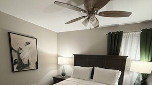 A neatly arranged bedroom with a bed, two bedside tables, a ceiling fan, a painting on the wall, and a window with curtains. - Relax at Cricket & Clover Retreat Near SU, LSU, Airport, Downtown, WiFi & AC (Baton Rouge)