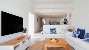 A modern living room with a flat-screen TV, white sofas with blue and white cushions, a wooden coffee table with fruit, and a kitchen area in the background. - Bright oceanfront bungalow with views, garden and coastal style, 91 (San Miguel de Abona)