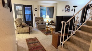 A living room with a sofa, armchair, and a TV. There is a staircase with a railing leading to an upper floor. - The most affordable winter adventure stay in McCall!  (McCall)