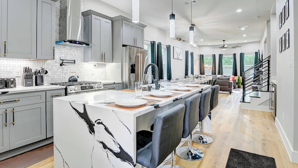 A modern kitchen with grey cabinets, a central island with bar stools, stainless steel appliances, pendant lighting, and a staircase leading to a dining area.