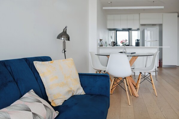 Ein modernes Wohnzimmer mit einem blauen Sofa, einem Esstisch mit weißen Stühlen und einer Küche im Hintergrund.
