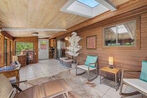 A cozy cabin interior with a wooden ceiling, a dining table, a sofa, a rocking chair, and a small side table with a lamp. - Lorel’s-Lake Area; Fenced for PETS; Mins to Downtown Rogers-Marina-Bike Trails (Little Flock)