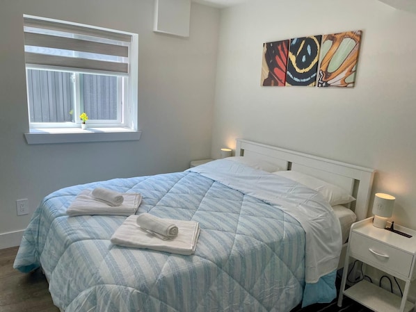 A neatly made bed with a striped duvet, a bedside table with a lamp, a window with curtains, and a wall with abstract art. - Garden Suite with Optional Interconnecting Suite, Save on Weekly & Monthly Stays (New Westminster)