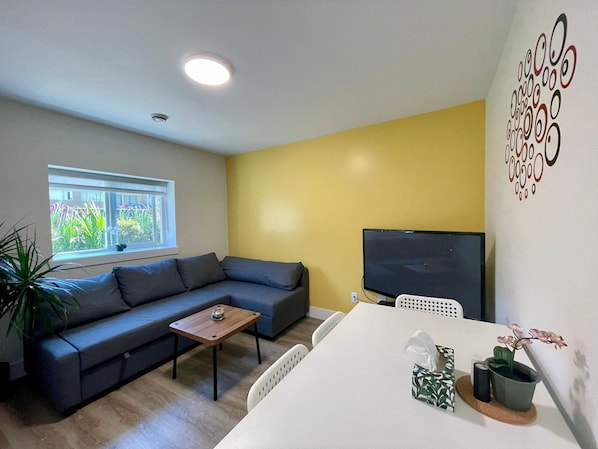 En moderne stue med en grå sofa, et lille træbord, et hvidt bord og et fjernsyn.