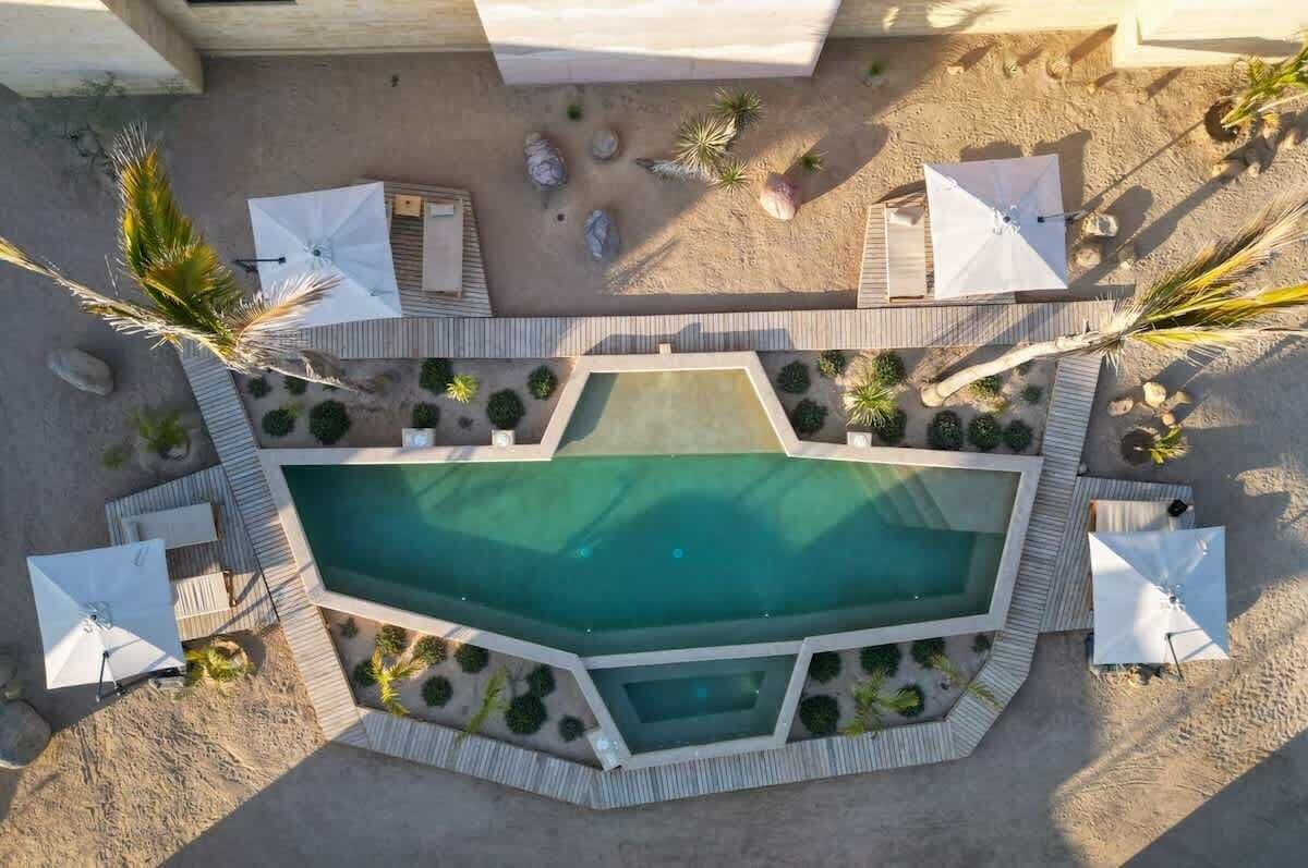 Vista aérea de una zona de piscina con una pileta rectangular, rodeada por una terraza de madera, sombrillas blancas y palmeras.