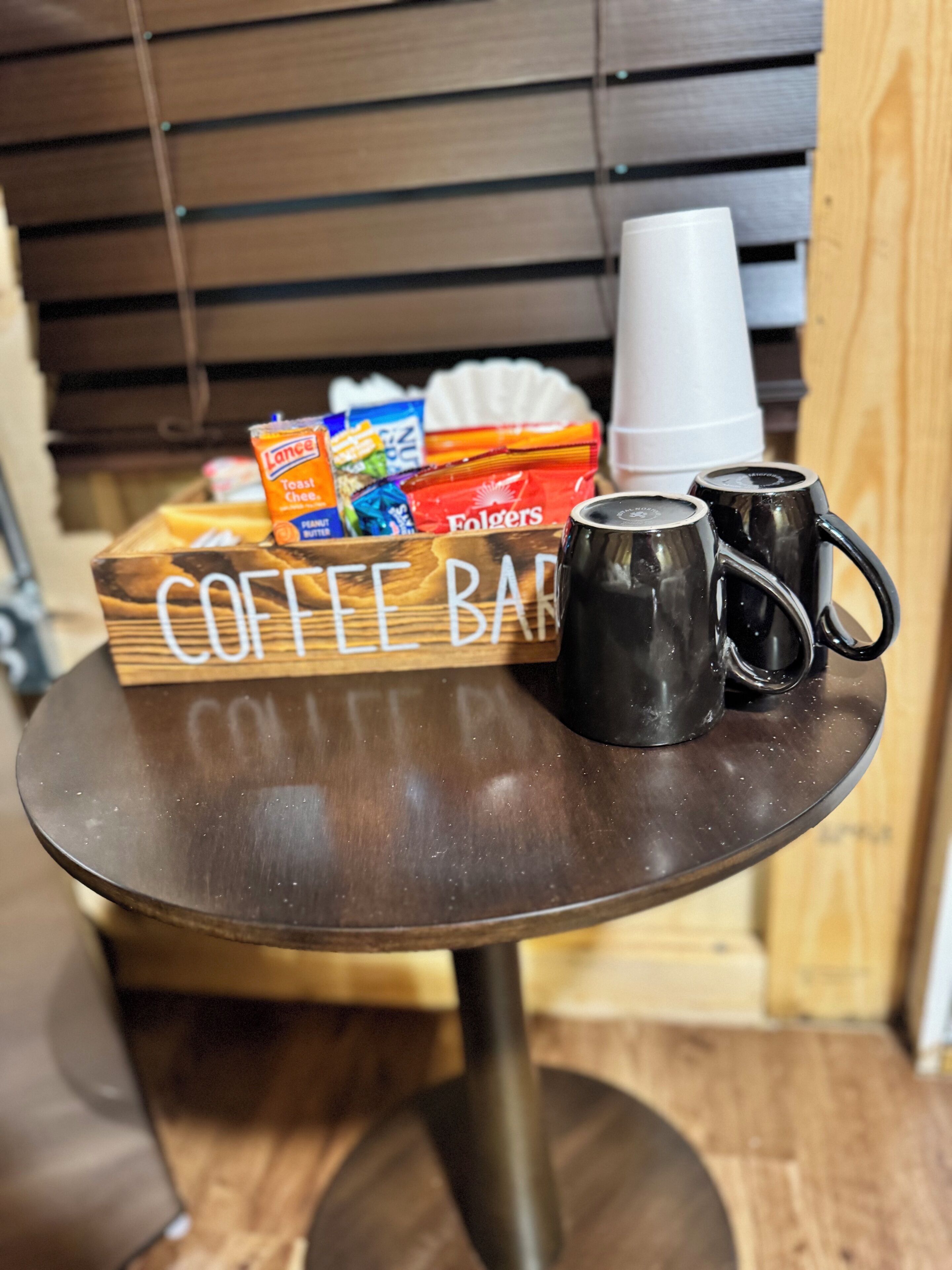 A coffee bar with a wooden sign, two black mugs, a white cup, and snack packages.
