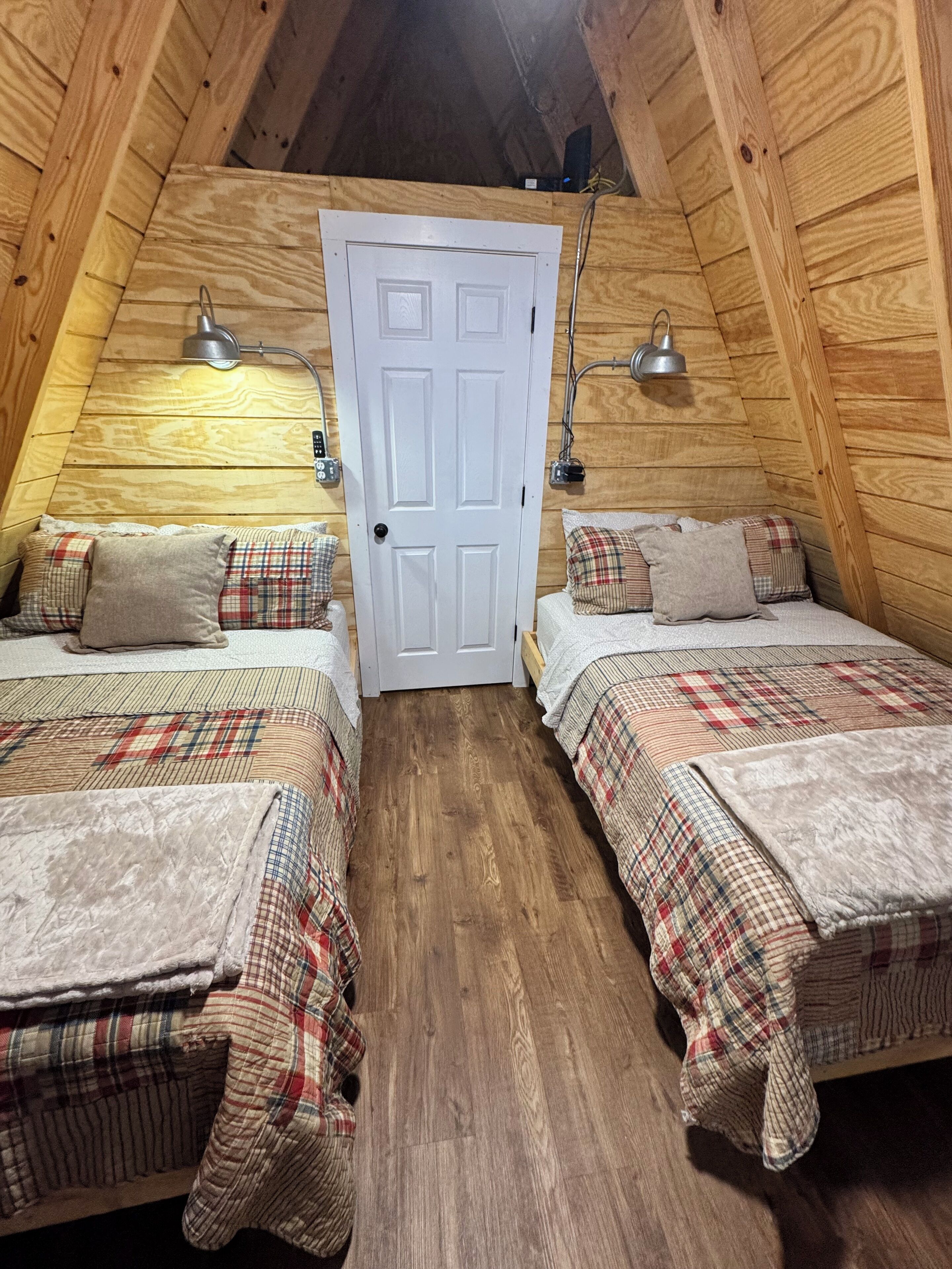 A cozy attic bedroom with two beds, plaid bedding, wooden walls, and a white door.