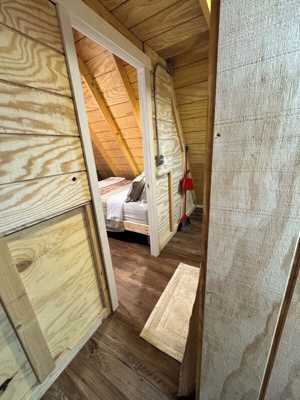 Un dormitorio rústico en el ático, con techo inclinado, una cama con almohadas, una escoba roja y una pequeña alfombra en el suelo.