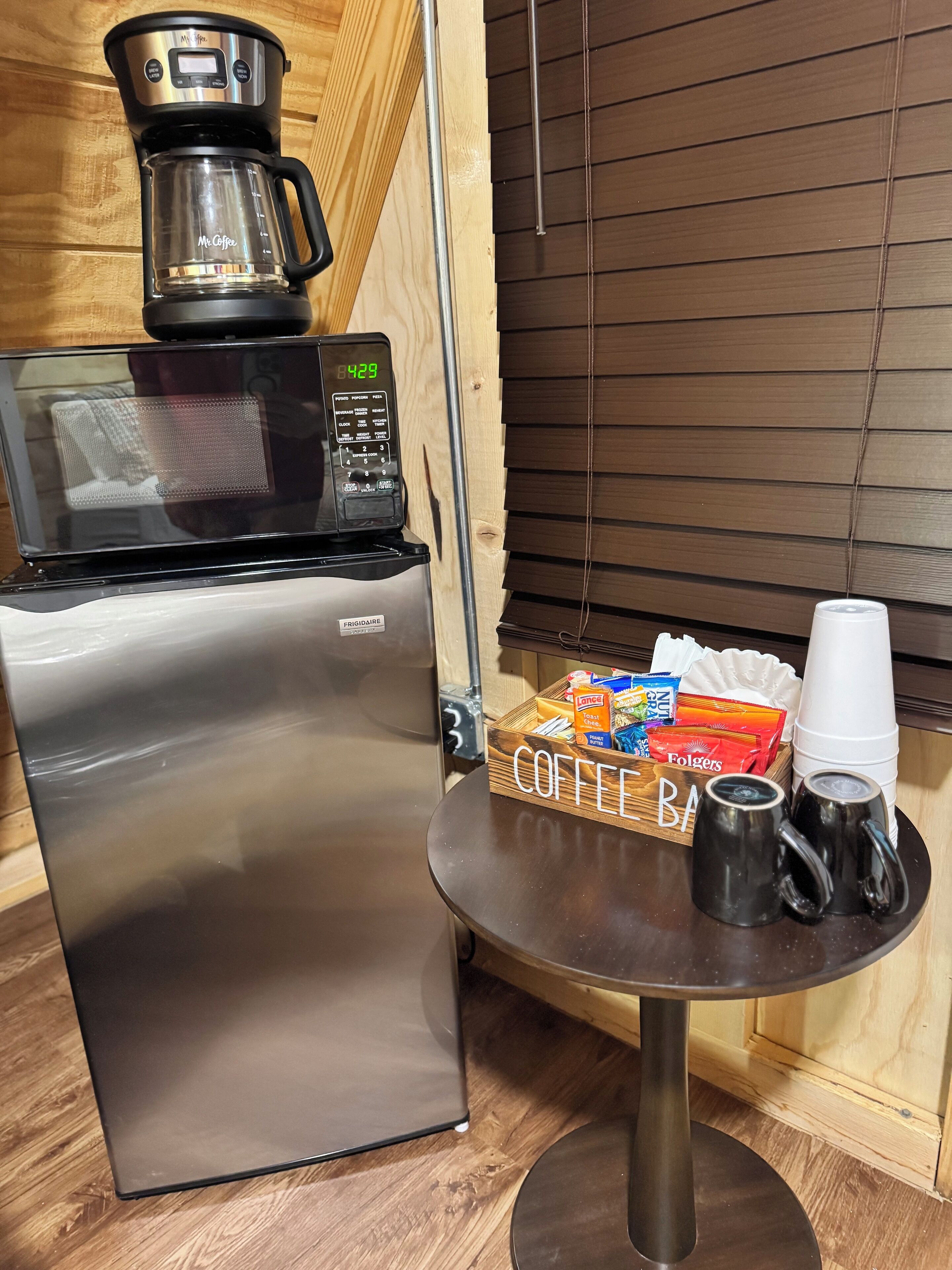 Una kitchenette compacta con refrigerador de acero inoxidable, cafetera, microondas y una pequeña mesa con tazas de café y una caja de café Folgers.