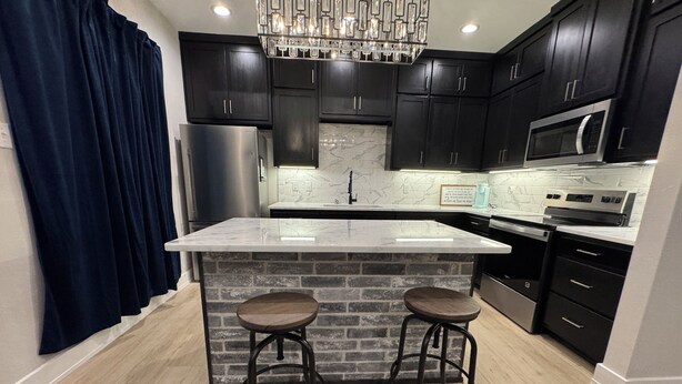 A modern kitchen with dark cabinetry, a marble island, stainless steel appliances, and a brick accent wall.